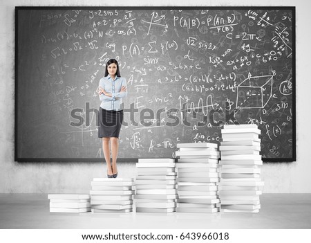 Ambitious blackhaired woman is standing on piles of books in a classroom with formulas written on a blackboard. Concept of woman in science