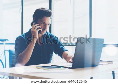 Elegant businessman working at sunny work place on laptop while sitting at the wooden table.Man using smartphone and analyze document on computer notebook.Blurred background.Horizontal