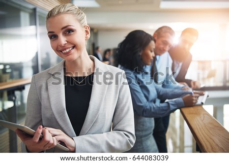 Smiling young blond business woman looking at camera and browsing tablet standing in office.