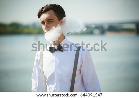young skinny man, elegantly stylishly dressed in white shirt, trousers with suspenders and bow tie. vaper guy Smokes an e-cigarette, releas vapors of smoke, against background of river and bridge