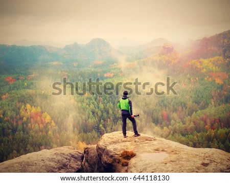 Amateur photographer prepare camera to takes impressive photos of  misty fall mountains. Tourist photographer at sharp rocky edge on high view point, colorful misty forest bellow.