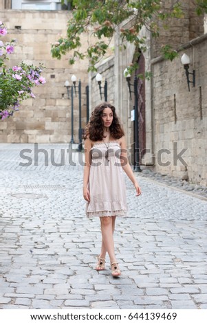 Full length portrait of curlie young girl wearing biege romantic dress strolling through old streets of Inner City of Baku and looking away from camera