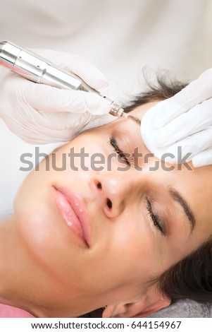 Close-up portrait of woman during making permanent makeup of eyebrows laying with eyes closed while cosmetologist in white gloves holding the needle.