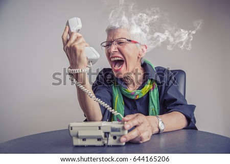 Angry, enraged senior woman yelling at a landline office phone, unhappy with customer service provided by the agent on the other side, giving off steam and smoke Royalty-Free Stock Photo #644165206
