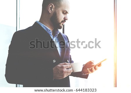 Man professional bookkeeper is holding cup of coffee and searching information on web site via cell telephone, while is standing near skyscraper window background with copy space for your advertising