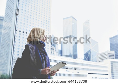 Young charming female tourist is holding digital table, while is standing in modern interior against big window with view of a business district background with copy space for your advertising content