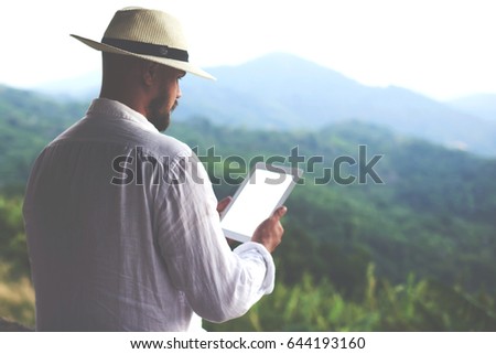 Hipster guy is holding digital tablet with copy space on the screen for your advertising text message or promotional content. Young man tourist is using touch pad,while is standing against jungle view