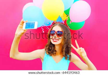 Portrait of happy smiling woman in birthday cap taking selfie picture by smartphone on a colorful balloons pink background