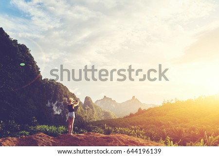 Hipster girl is shooting video of beautiful sundown on cell telephone, while is standing against Amazon scenery background with copy space. Female tourist is taking photo with mobile phone camera