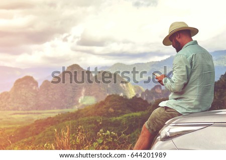 Man tourist is searching way in navigation via mobile phone, while is taking break between road trip on suv. Young hipster guy is watching photos on cell telephone, while is sitting on a car hood  