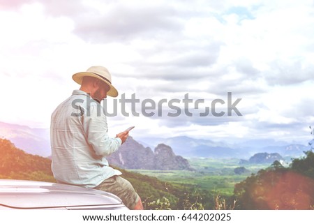 Young man is looking way on navigation via cell telephone during his amazing summer adventure in Thailand. Bearded male is watching photos on smart phone, while is sitting on a suv hood against jungle