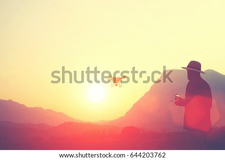 Man wanderer shooting video with radio controlled drone, while is standing in beautiful evening against high mountain and sunset. Male is using flying quadcopter for make amazing pictures from above