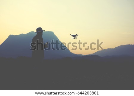 Silhouette of a blog author is taking photo of beautiful view with flying quadcopter for his internet page. Young male is shooting video on modern RC multirotor, while standing in evening against alp 