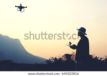 Silhouette of a man photographer is taking photo on flying quadcopter with camera, while is standing against sunset and alp. Young male traveler is using RC multicopter during summer trip overseas