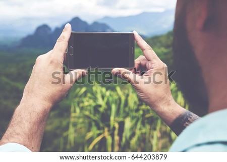 Back view of bearded man is taking photo with cell telephone camera,while is standing on mountain with subtropical forest. Young hipster guy is shooting video on smart phone of a amazing wild nature
