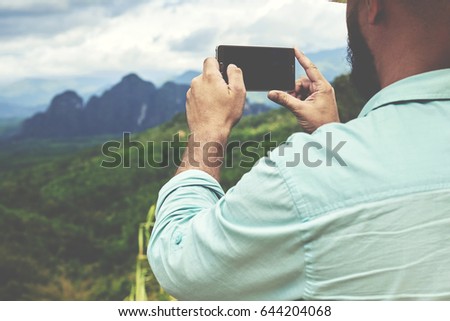 Back view of a young man traveler is taking photo with cell telephone camera of a beautiful jungle landscape. Young male wanderer is shooting video on mobile phone during summer adventure in Asia 