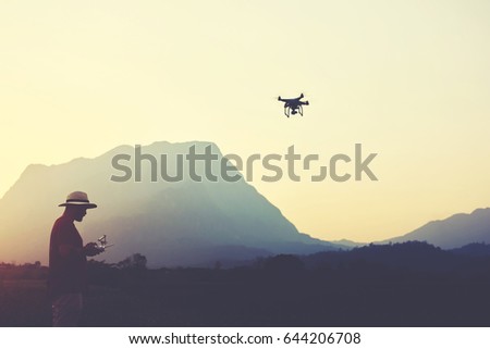 Young man traveler is shooting video on multirotor, while is standing in rural against high mountains in evening. Male wanderer is using radio-controlled quadcopter during his summer weekend abroad