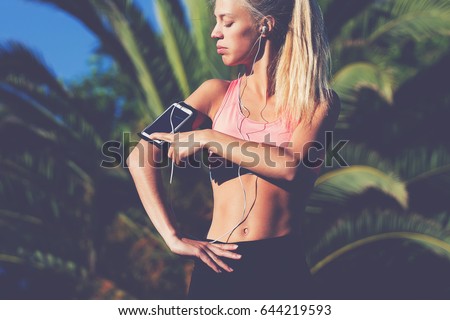 Half length portrait of athletic female touching screen of smart phone in running armband while listening music in headphones, fit woman taking break after workout outdoors in sunny summer day