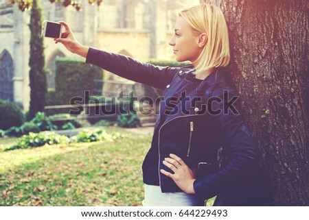 Stylish hipster girl is making self portrait with mobile phone camera, while is standing outdoors near old tree. Attractive woman traveler is photographing herself on cell telephone during her walking