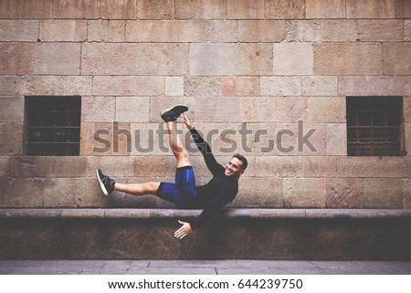 Handsome male athlete doing exercising for legs while lying on a concrete bench, smiling sportsman doing workout outdoors against brick wall with copy space area for text message or content