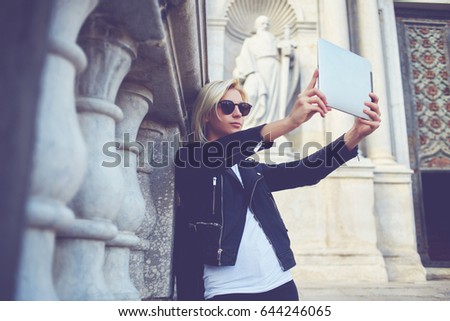 Stylish woman in sunglasses is making self portrait with front touch pad camera on background of old church.Fashionable female is photographing herself on digital tablet, while standing outside museum