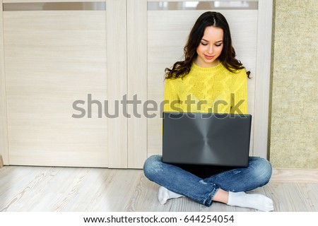 Beautiful young woman sits on the floor with a laptop in her lap. Looks at the screen and communicates in social networks.