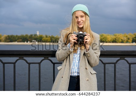 Positive attractive young female photographer amateur enjoying spending time on hobby strolling outdoors with vintage camera, portrait of hipster girl taking pictures of scenery standing on bridge

