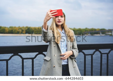 Charming teenaged blonde hipster girl dressed in casual wear making selfie for updating profile picture, attractive blogger making streaming video standing on bridge near copy space for advertising