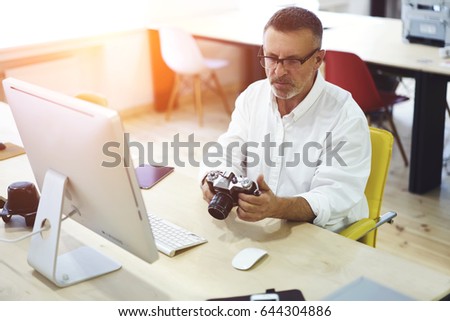 Male programmer 50 years old looking picture made by colleague about modern design of interior during free time. Mature professional it developer using vintage camera for work process sitting indoors
