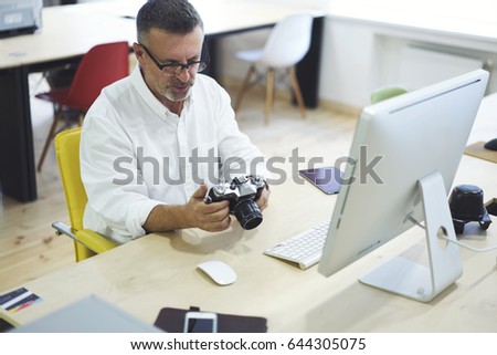 Mature experienced architector in eyeglasses looking photos from building office planning on vintage camera for understanding and developing future project sitting on workplace at modern computer 