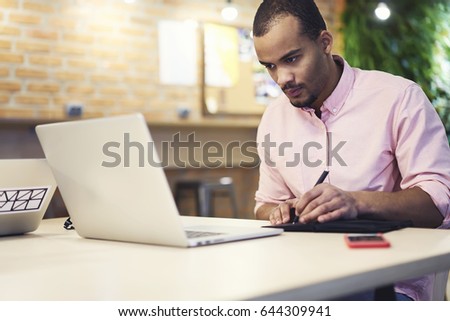 Skilled concentrated afro american graphic designer drawing sketch of creative planning in notebook while watching online scheme of office building on modern laptop computer sitting in office
