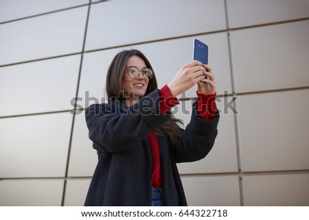 Good looking cheerful woman dressed stylish overcoat making picture using application on modern telephone connected to wireless internet and sharing image for friends in social network while strolling