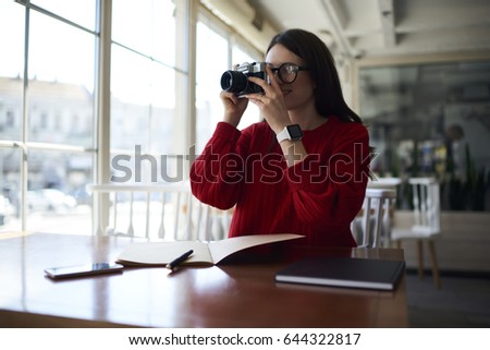 Female student taking pictures for studying project sitting in coffee shop with notepad for records.Young woman making photos on vintage camera while writing some notes in diary during free time 