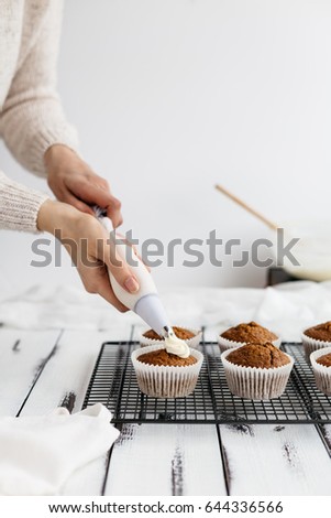 Woman Piping Cream Cheese Icing onto Freshly Baked Carrot Cupcakes on White Wooden Table 