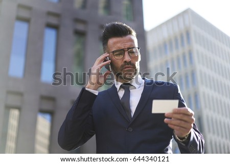 Prosperous mature business man having phone conversation with banking service using contact of call center from blanked visit card, professional male owner of company contact with partner via mobile
