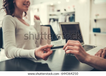 Attractive girl is giving a credit card to shop assistant and smiling while doing shopping in boutique Royalty-Free Stock Photo #644364250