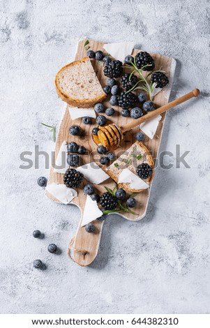 Berries blackberry and blueberry, honey on dipper, rosemary, sliced goat cheese with bread served on wooden board over gray texture background. Summer sandwich. Top view with space