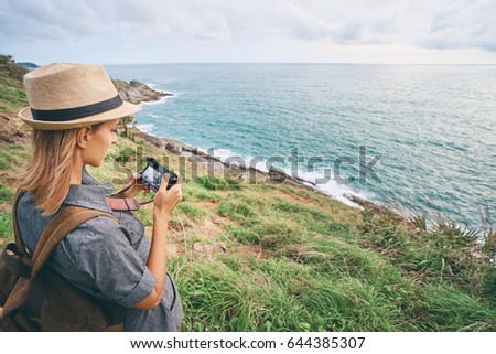 Photography and travel. Young woman in hat with rucksack holding camera enjoying sea view.