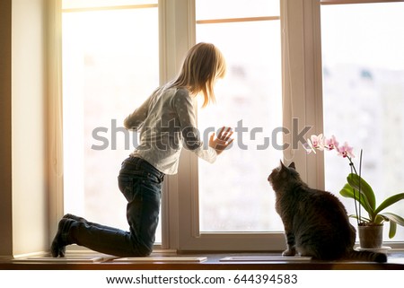 The girl is kneeling a window sill and looks out of the window. The cat costs on a window sill near the girl