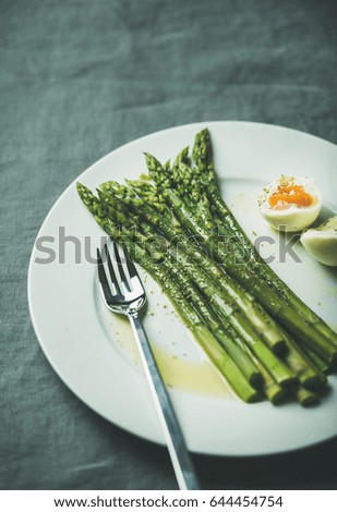 Cooked asparagus with soft-boiled egg and herbs on white round plate over grey textile background, selective focus, copy space. Clean eating food concept