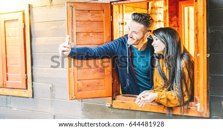 Loving couple taking a selfie outside the window of their wooden house - Young lovers taking pictures with mobile phone camera - Selfie addiction - Soft focus on female face