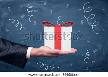 A businessman's hand turned up and a small gift box with a red bow standing on chalkboard background. Promotions and bonuses. Getting clients interested. Best offers.