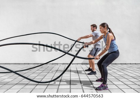 Fitness people exercising with battle ropes at gym. Woman and man couple training together doing battling rope workout working out arms and cardio for cross fit exercises. Royalty-Free Stock Photo #644570563