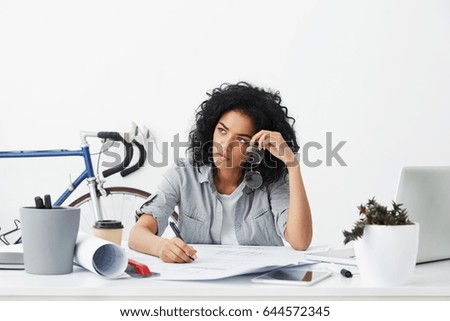 Overwork concept. Portrait of unhappy and tired young Afro American female architect working on blueprints at her workplace having bored look, holding pen in one hand and eyeglasses in other