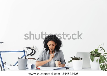 Fashionable female Afro American student doing her homework designing new buildings sitting in cosy home interior drinking coffee holding pen and having thoughtful expression. People and education