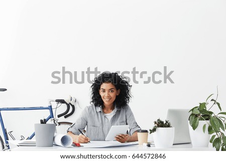 Indoor shot of attractive young African student architect working at her project work holding pen and tablet in her hands having bicycle behind her back going to ride it and to relax after hard work.