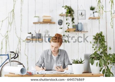 Hardworking concentrated redhead Caucasian female engineer busy filling in technical specifications sitting at her workspace using mobile phone against cozy light office interior background
