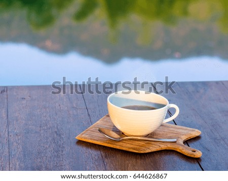 Hot coffee on wooden saucer and on wooden table with reflection of mountain and tree in water under morning sunlight 