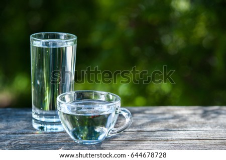 Glass and a cup with transparent, cool clean water on a green garden background. Clean water, health, food industry, health, Ecology, environmental protection, thirst quenching 