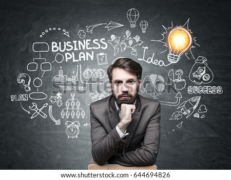 Portrait of a serious bearded businessman wearing a brown suit and sitting near a blackboard with a business plan on it